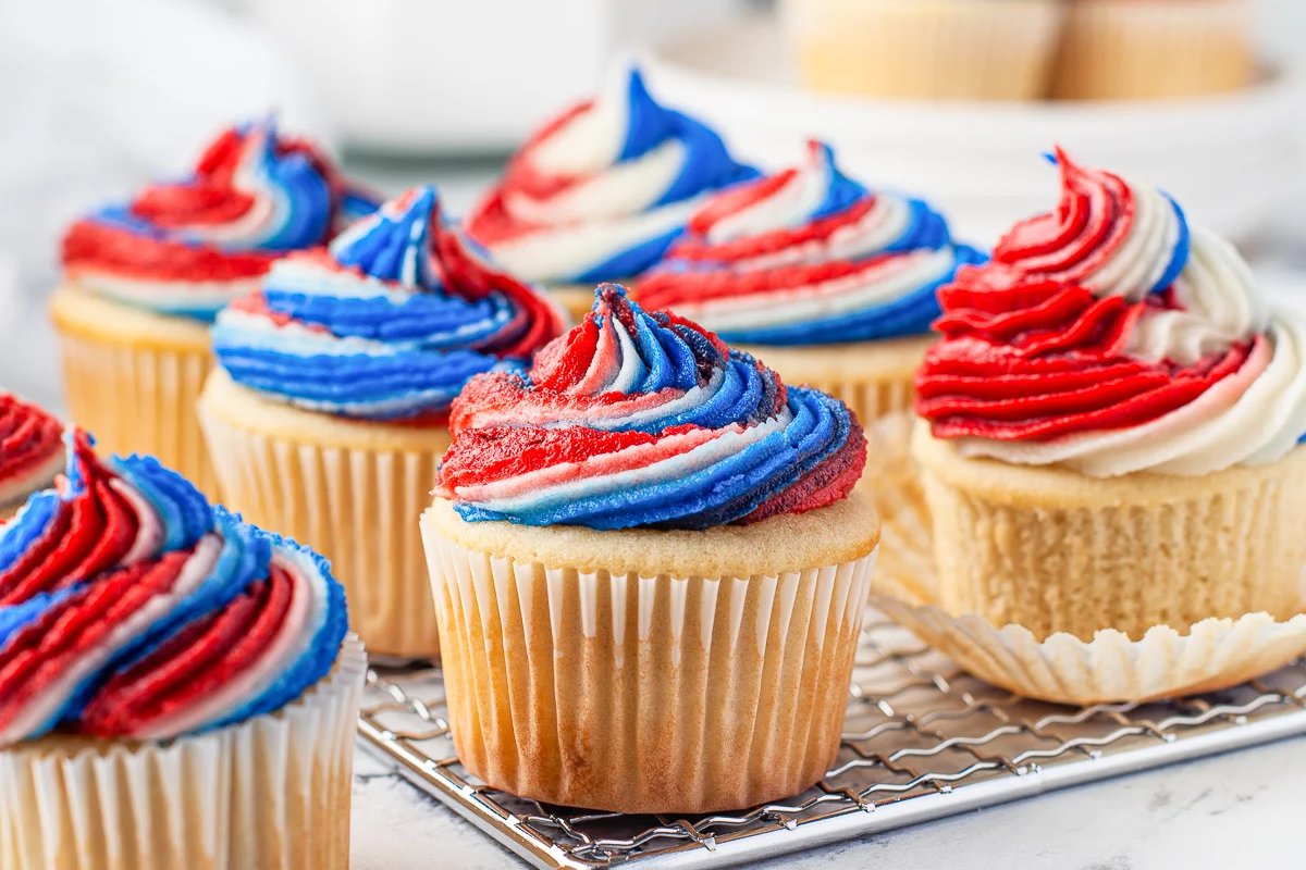 Patriotic desserts line the cooling rack, featuring cupcakes with red, white, and blue swirled frosting. More festive treats can be seen in the background, completing the celebratory display.