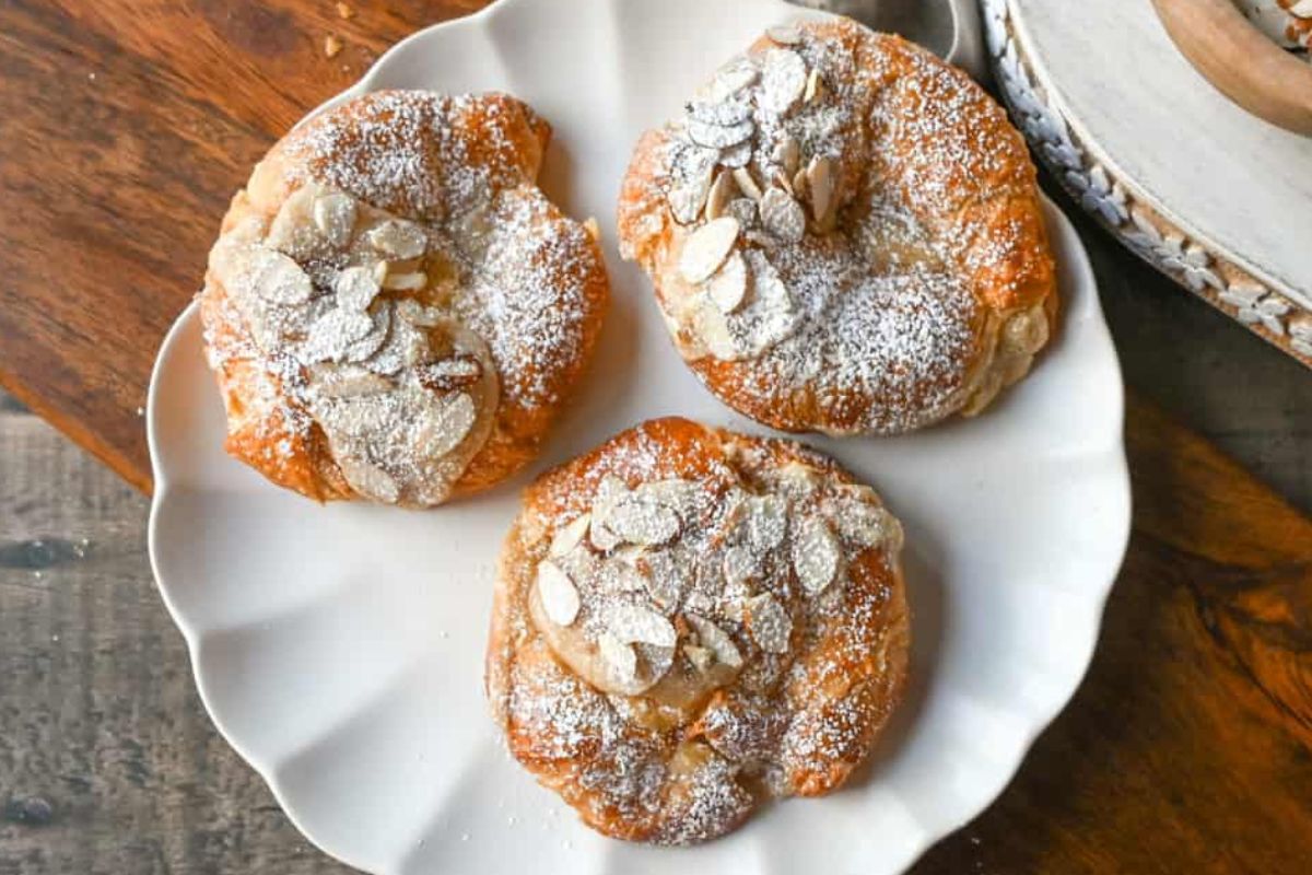 Three almond pastries topped with powdered sugar and sliced almonds on a white plate, these shortcut desserts are perfect for a quick yet delightful treat.