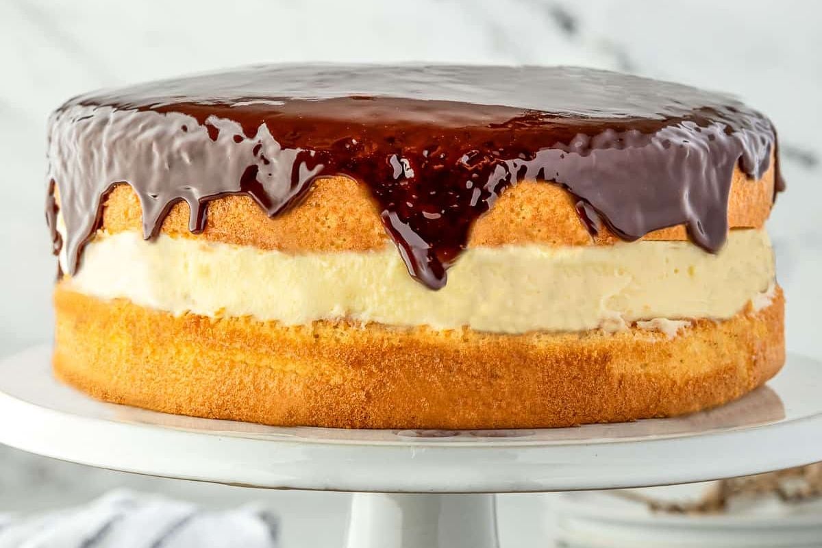 A layered sponge cake with a cream filling and chocolate glaze on top, displayed on a white cake stand—perfect for fans of shortcut desserts.