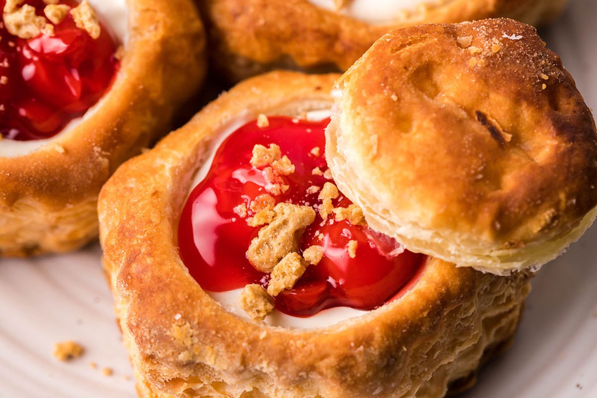 Close-up of pastries with golden-brown, flaky crusts filled with white cream and topped with red fruit filling and crumbled pieces. One pastry has its top slightly lifted.