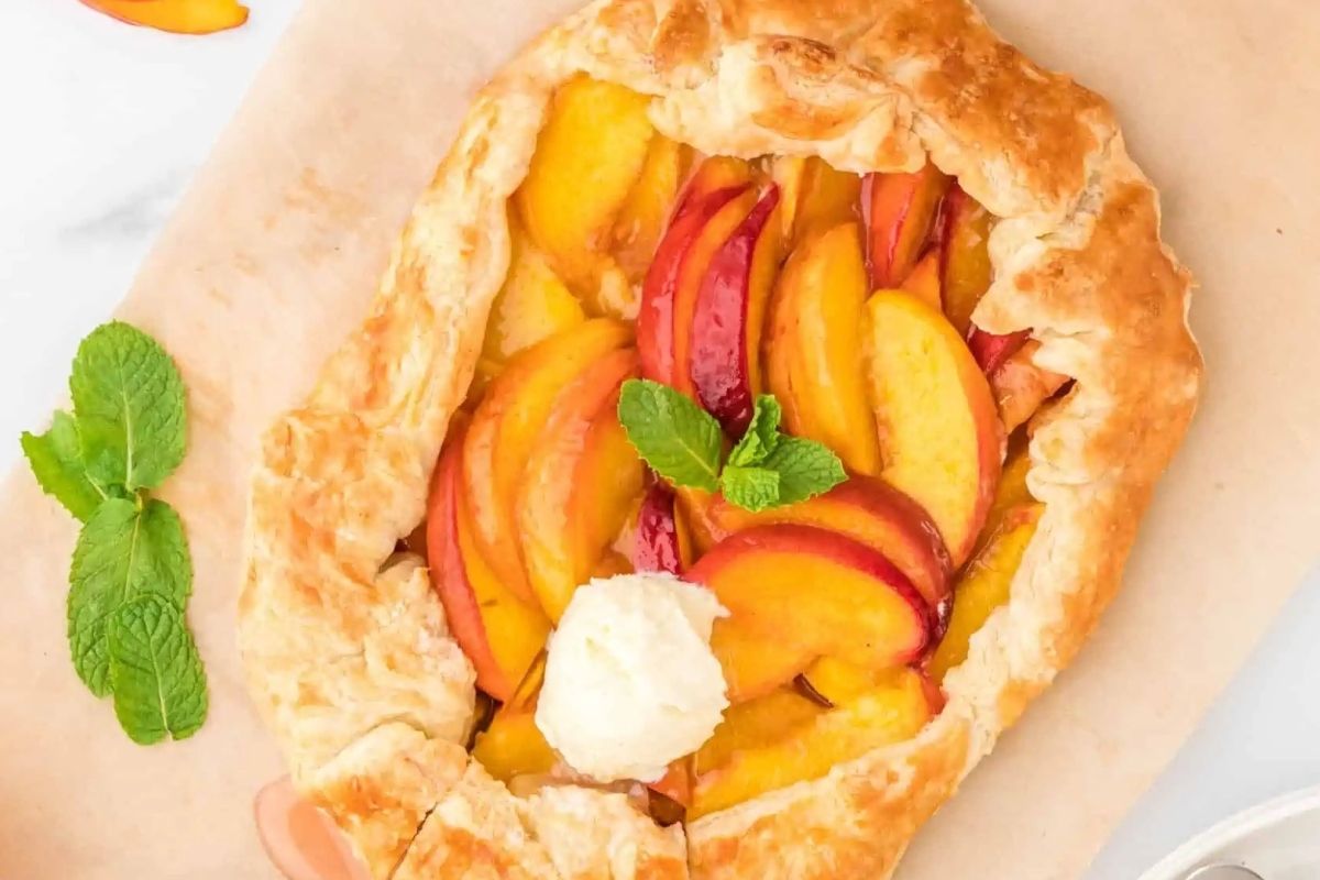A peach galette with a golden, flaky crust, topped with a scoop of vanilla ice cream and a sprig of mint, is placed on a piece of parchment paper&mdash;one of the most delightful and easy desserts to brighten your day.