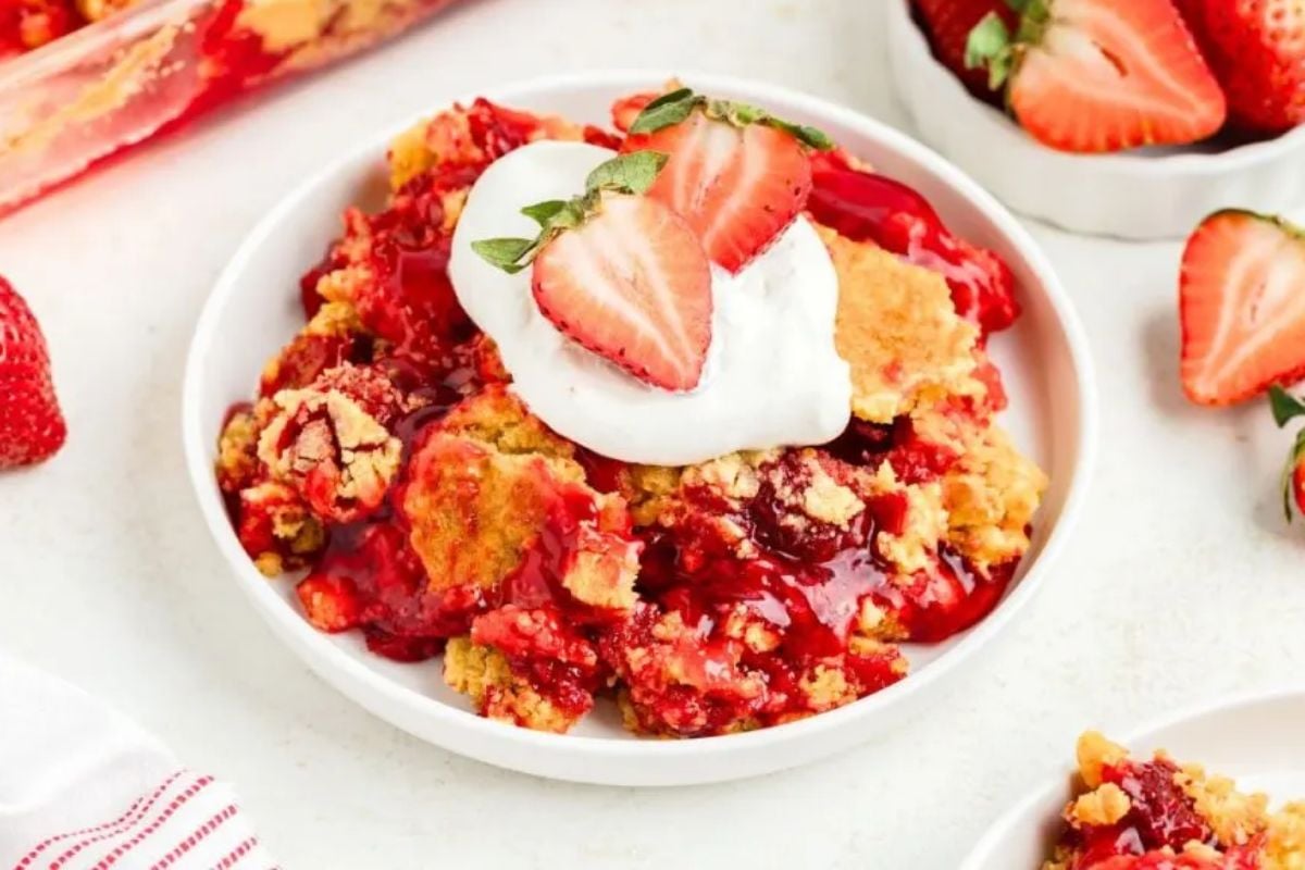 A white plate of strawberry dump cake topped with a dollop of whipped cream and fresh strawberry slices. A bowl of whole strawberries is visible in the background, showcasing an easy approach to shortcut desserts.