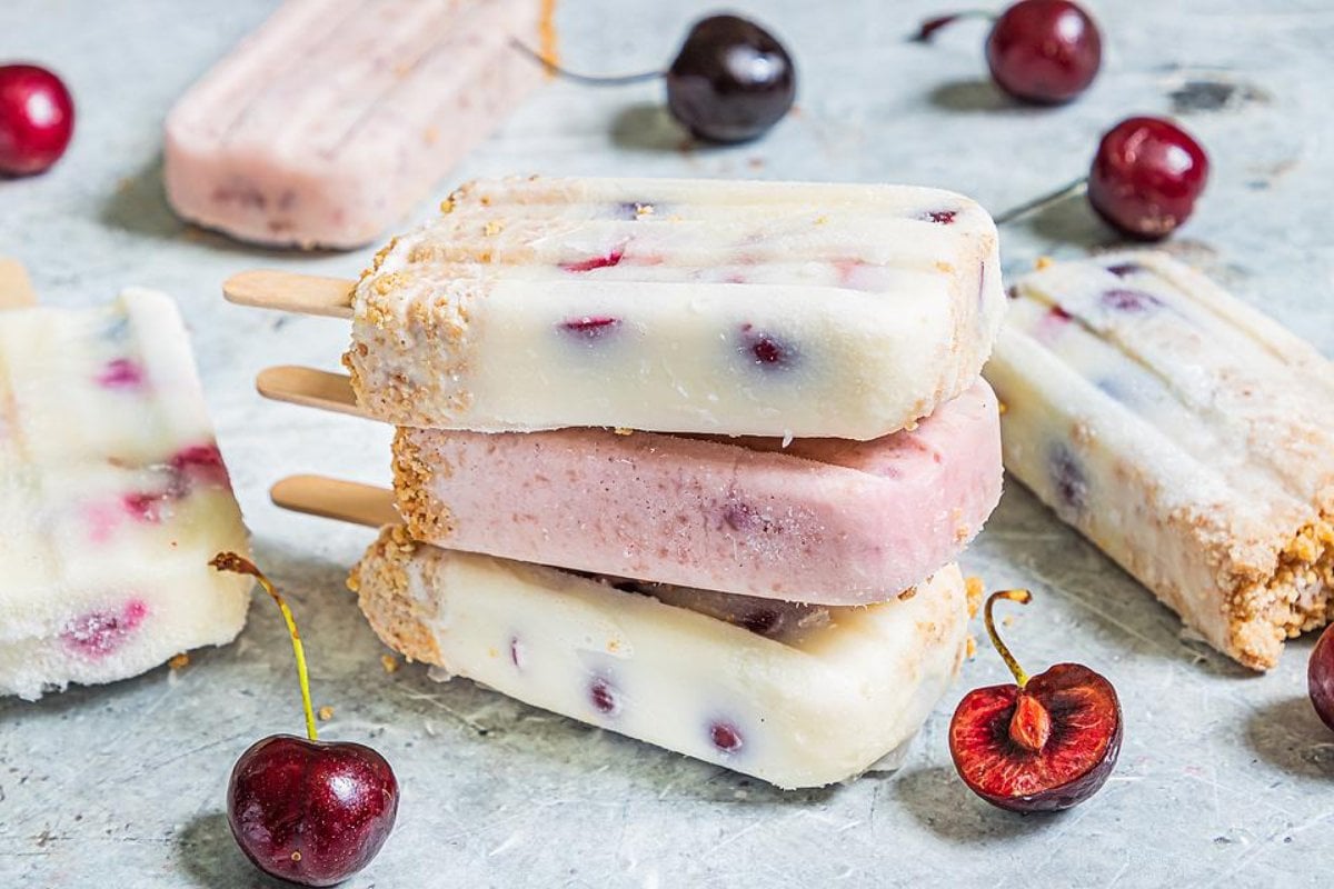 Assorted cherry popsicles are arranged on a light surface, surrounded by fresh cherries. Featuring different layers and colors, these treats highlight an array of flavors inspired by classic cherry recipes.