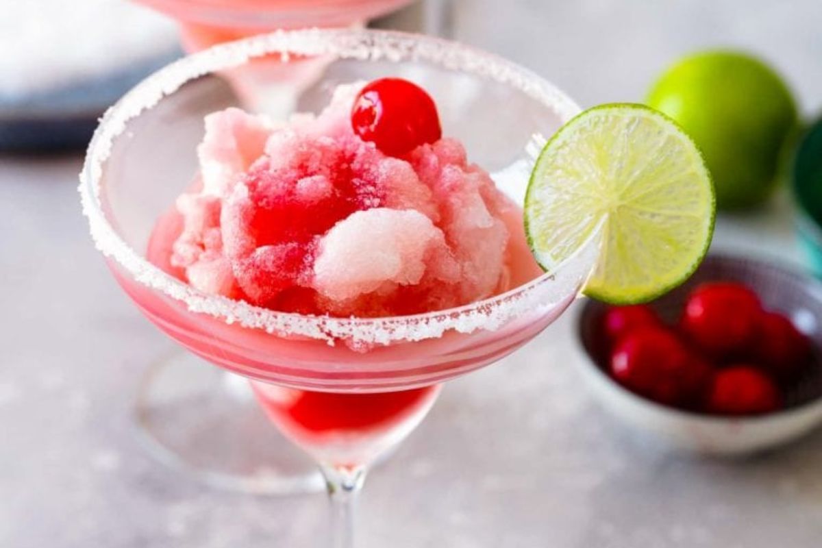 A glass with pink slush, garnished with a cherry and a lime slice, sits on a surface near a bowl of red cherries and a whole lime, showcasing one of the best cherry recipes for summertime refreshment.
