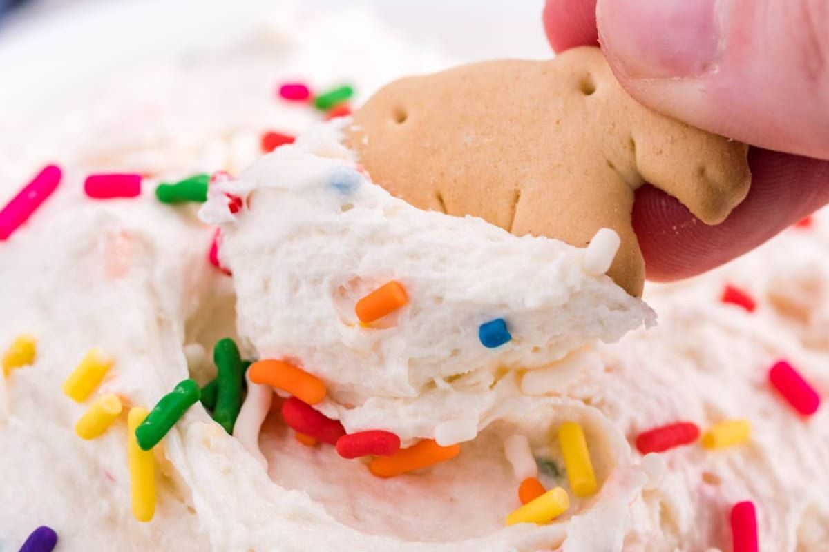 A hand dips an animal cracker into creamy dip with colorful sprinkles—a playful twist inspired by cool whip recipes.