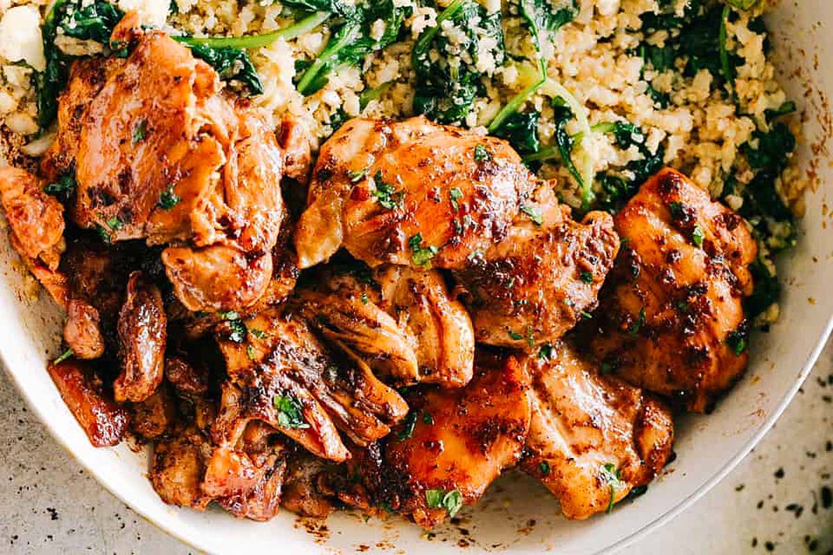 Grilled chicken thigh pieces and sautéed spinach served with crumbled cauliflower rice in a white bowl.