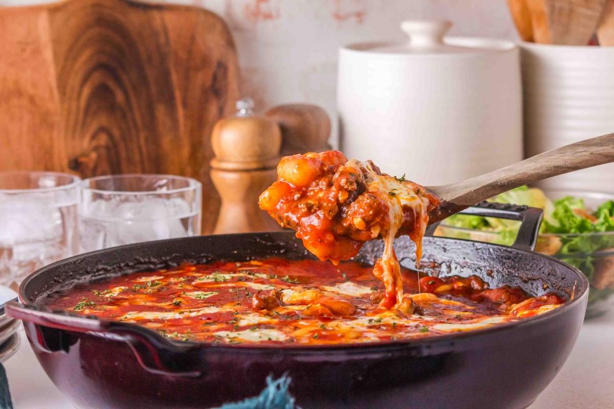 A wooden spoon lifts cheesy, baked gnocchi in tomato sauce from a red skillet on a kitchen counter—an inviting choice for cozy baked dinners.