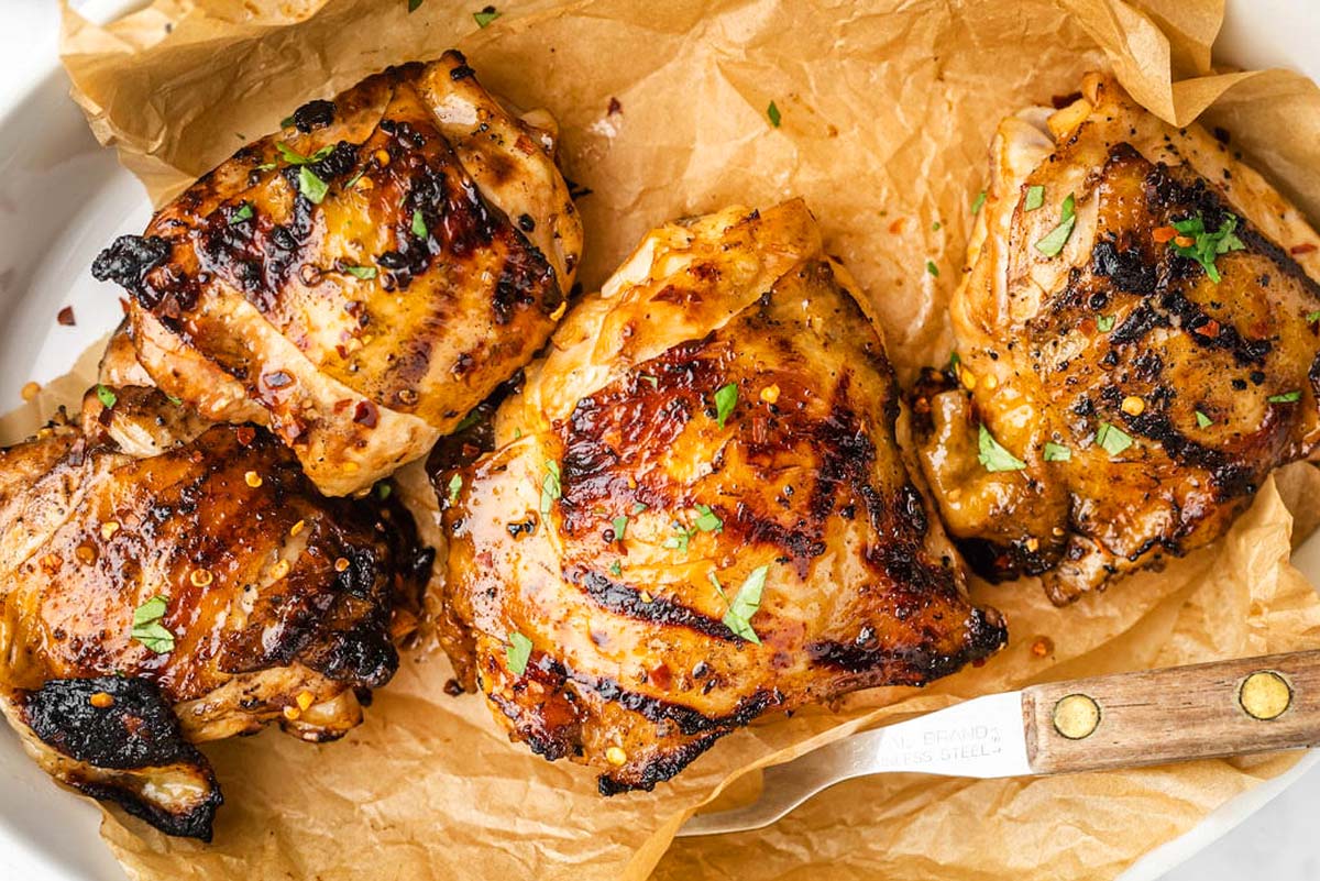 Four grilled chicken thighs, garnished with herbs, are placed on a sheet of parchment paper with a knife nearby—perfect for those seeking delicious chicken thigh recipes.