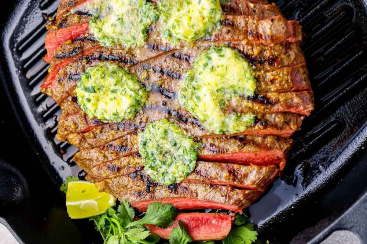A grilled flank steak with perfect grill marks, topped with dollops of herb butter, garnished with cilantro and lime wedges, on a black grill pan.