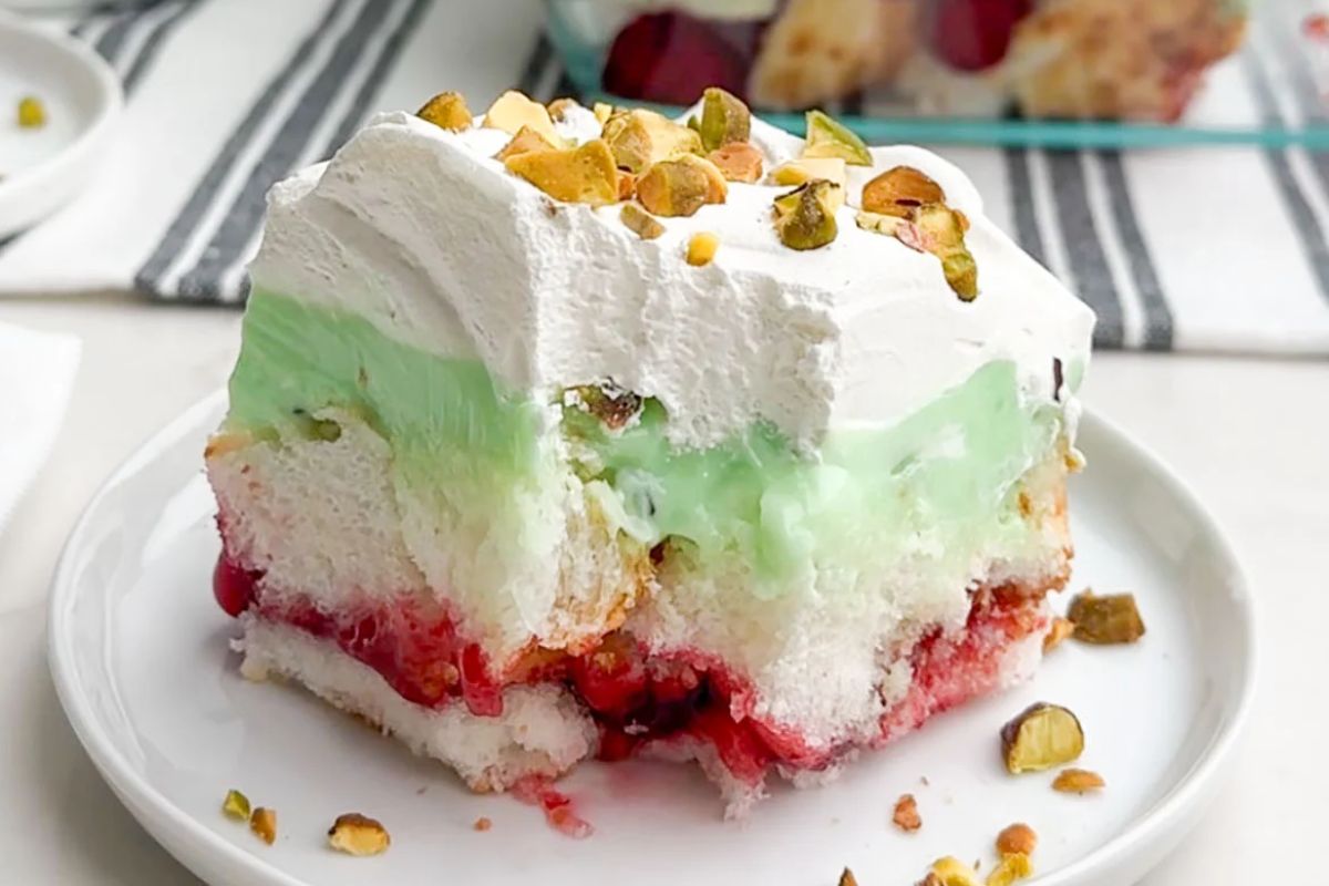 A slice of layered dessert with white cake, cherry fruit filling, green pudding, and whipped cream topped with chopped pistachios, served on a white plate is one of the finest cherry recipes you can try.