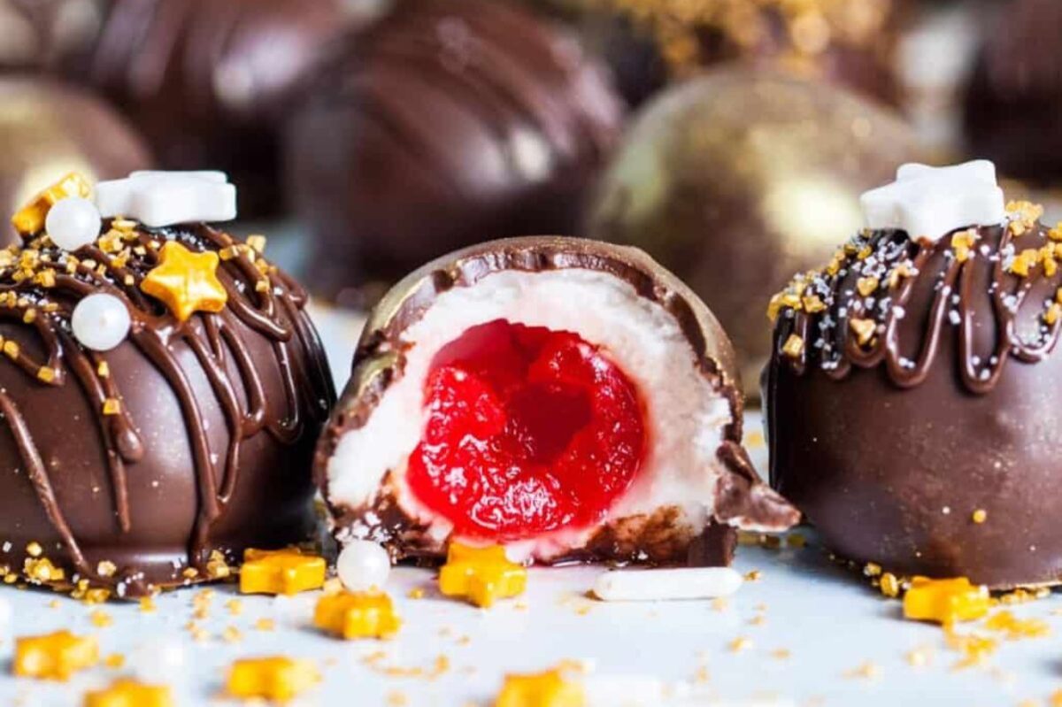 Several chocolate truffles with decorative sprinkles are shown. One truffle is cut in half, revealing a red and white cherry filling inside, reminiscent of classic cherry recipes.