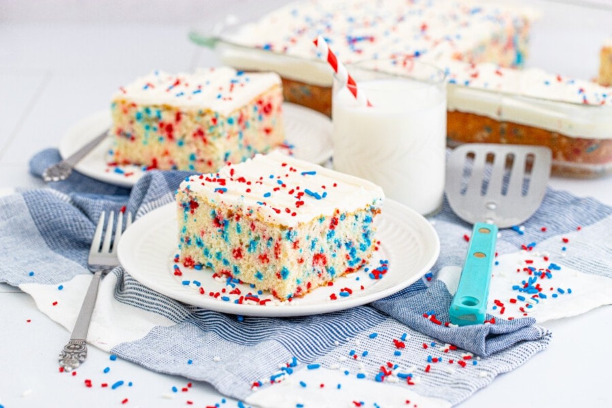 A slice of cake with white frosting and red and blue sprinkles on a plate, next to a glass of milk with striped straws. Another slice and the remaining cake in a pan are in the background&mdash;perfect examples of patriotic desserts.