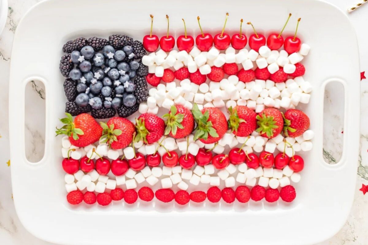 A rectangular tray with fruits and marshmallows arranged in the shape of the American flag. Patriotic desserts like these feature blueberries and blackberries forming the blue section, while cherries, strawberries, raspberries, and marshmallows create the red and white stripes.