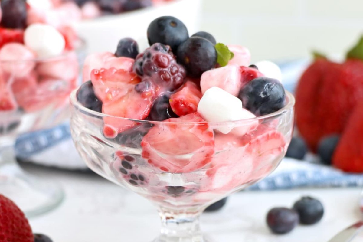 A glass dessert dish filled with a creamy fruit salad, including strawberries, blueberries, blackberries, and mini marshmallows makes for perfect patriotic desserts.
