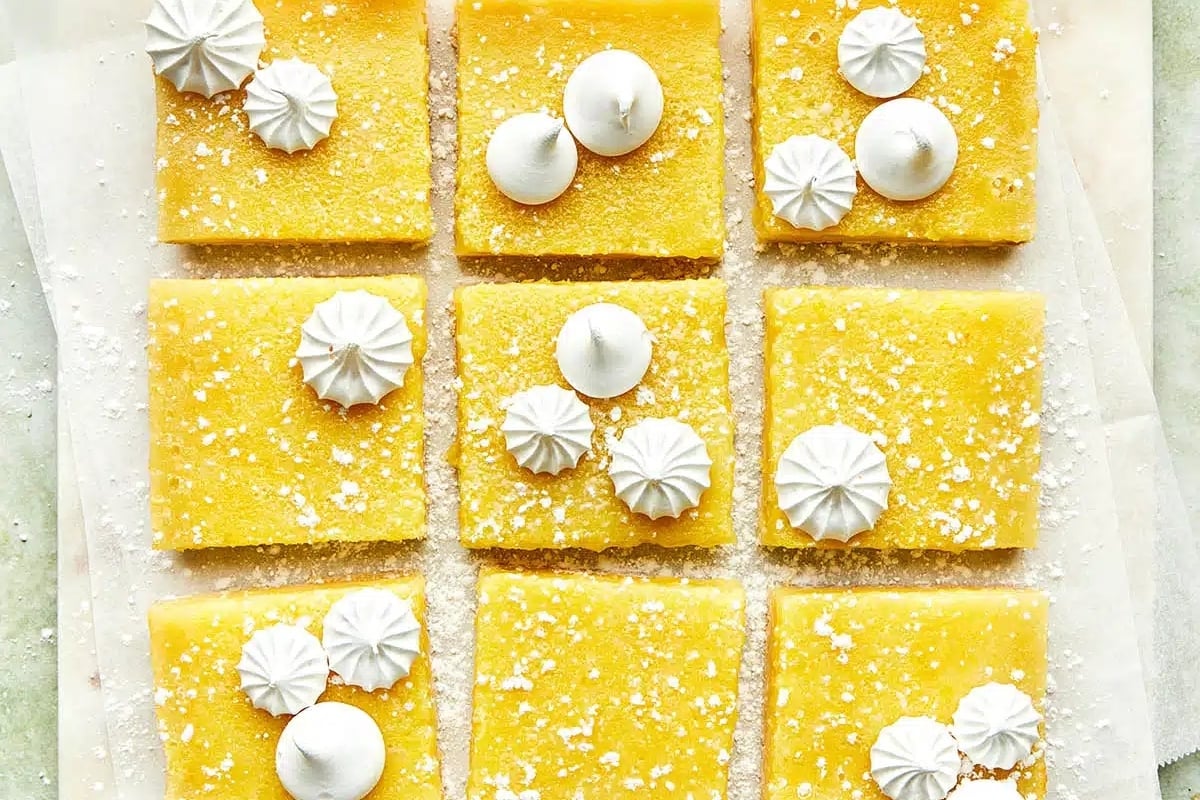 Nine lemon bars arranged in a grid, each topped with small meringues and lightly dusted with powdered sugar. These easy desserts make for a delightful treat at any gathering.