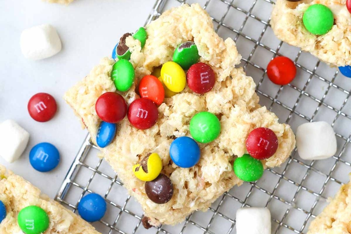 A close-up of a rice crispy treat topped with colorful chocolate candies, placed on a cooling rack. Small marshmallows and more candies are scattered around, making it one of the easiest shortcut desserts to whip up.