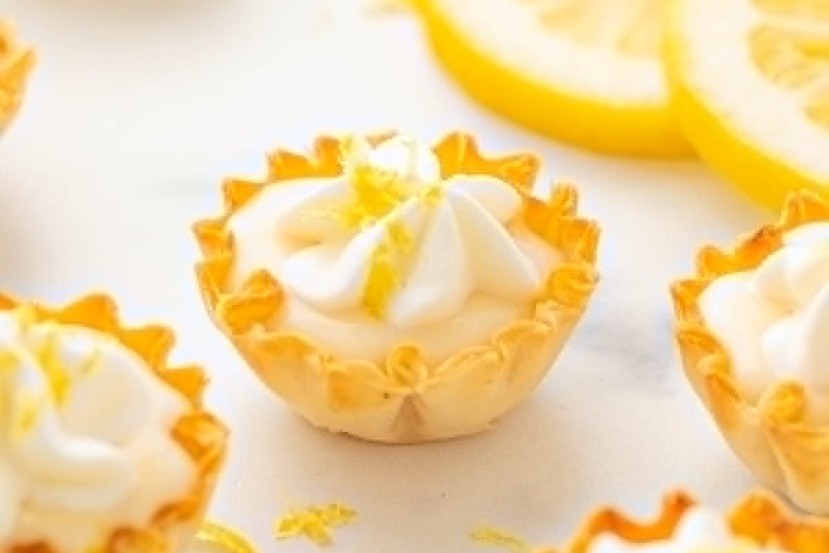A close-up of a lemon tart with a dollop of whipped cream on top, garnished with lemon zest. Slices of lemon are visible in the background, showcasing one of the ultimate shortcut desserts.