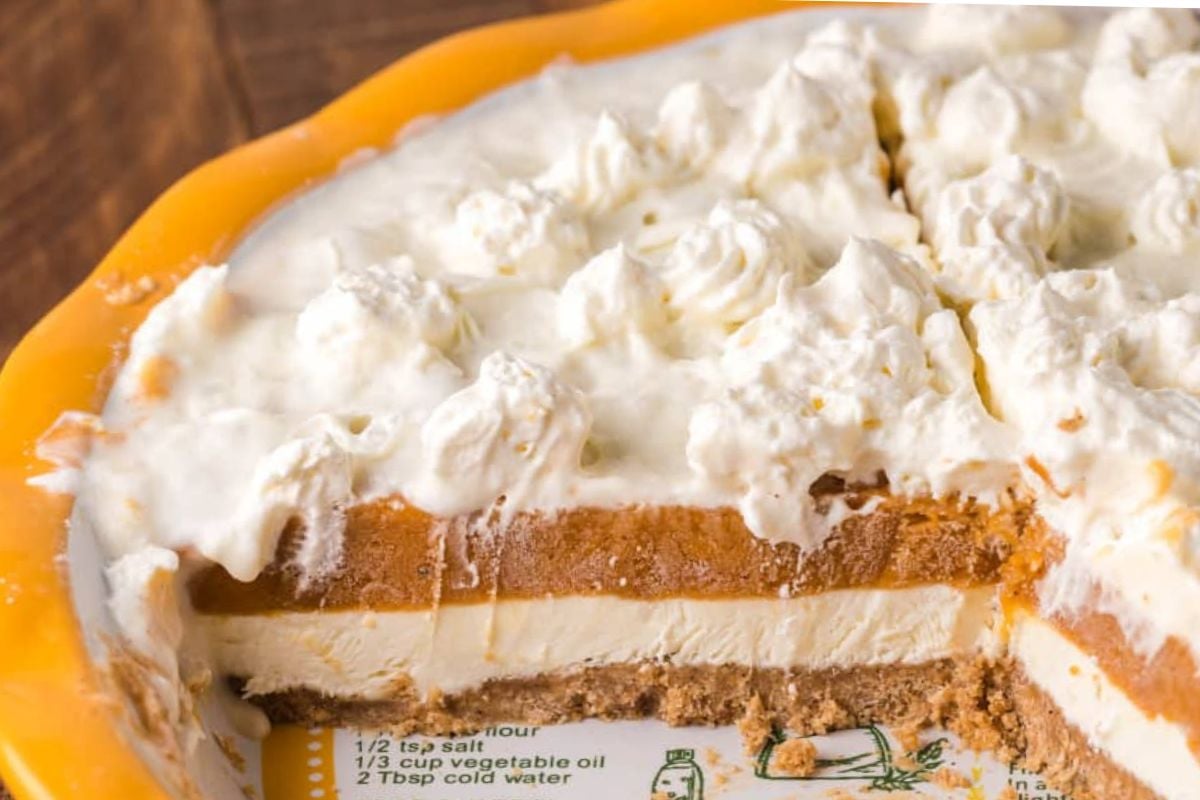 A pie with a graham cracker crust, layers of cream filling and pumpkin filling, topped with dollops of whipped cream on a serving dish with part of the pie missing.