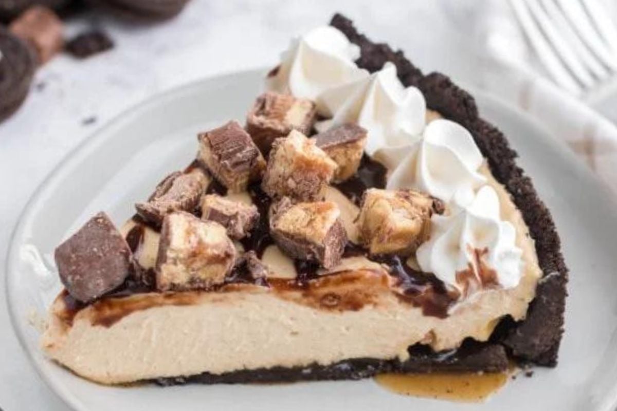 A slice of chocolate peanut butter pie topped with chopped peanut butter cups, whipped cream dollops, and chocolate drizzle on a white plate.