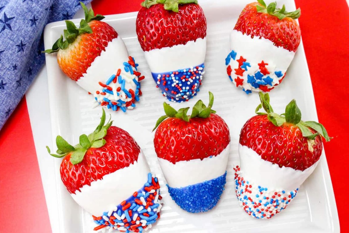 Patriotic desserts of strawberries dipped in red, white, and blue chocolate, decorated with various patriotic sprinkles, arranged on a white plate.