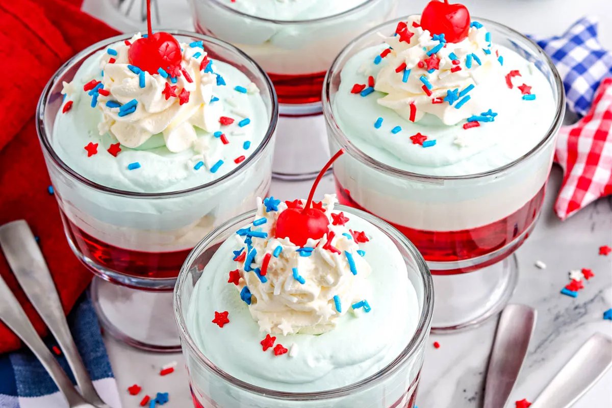 Four glass cups filled with layered red, white, and blue dessert, topped with whipped cream, colorful sprinkles, and a cherry on top are arranged on a marble surface with spoons and napkins. These patriotic desserts make for a festive and delicious treat.