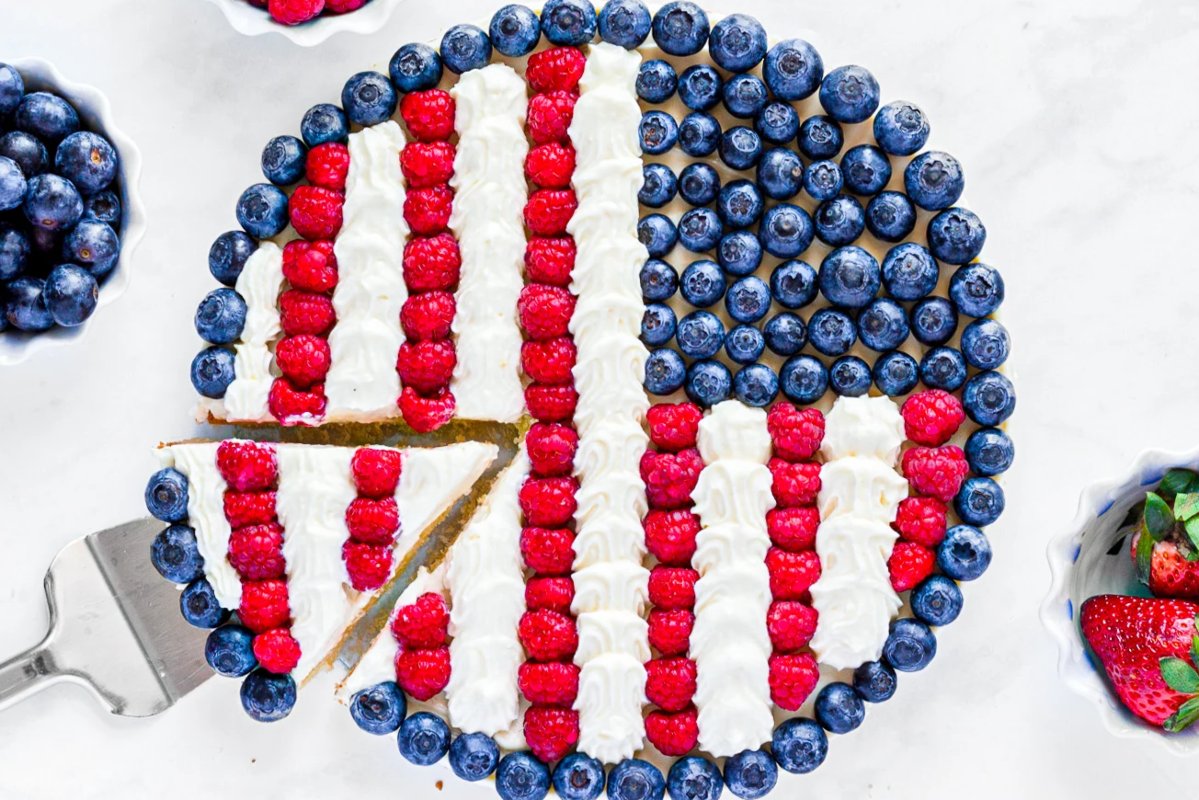 A round berry cake, one of the finest patriotic desserts, features patterns made of blueberries, raspberries, and white cream. A slice has been cut out and placed on a server. Bowls of berries surround the cake on a white surface.