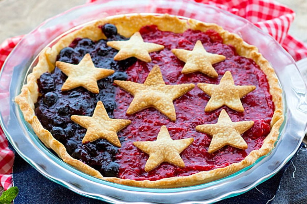 A patriotic dessert featuring a pie with a red and blue filling, topped with star-shaped dough cutouts, arranged in a glass pie dish on a red checkered cloth.