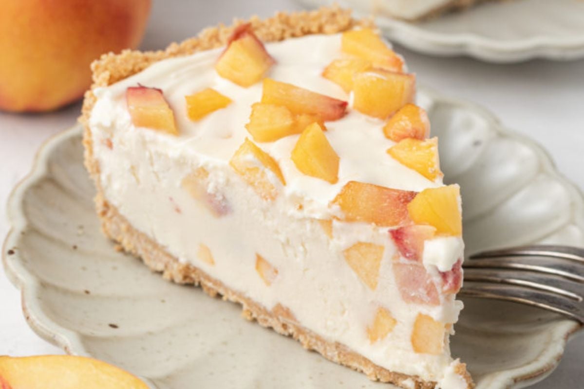 A slice of peach cheesecake on a light-colored plate with a fork. The cheesecake is topped and filled with pieces of fresh peach, making it one of those easy fruit desserts you can't resist. Another peach sits in the background, adding to the allure of this delightful treat.