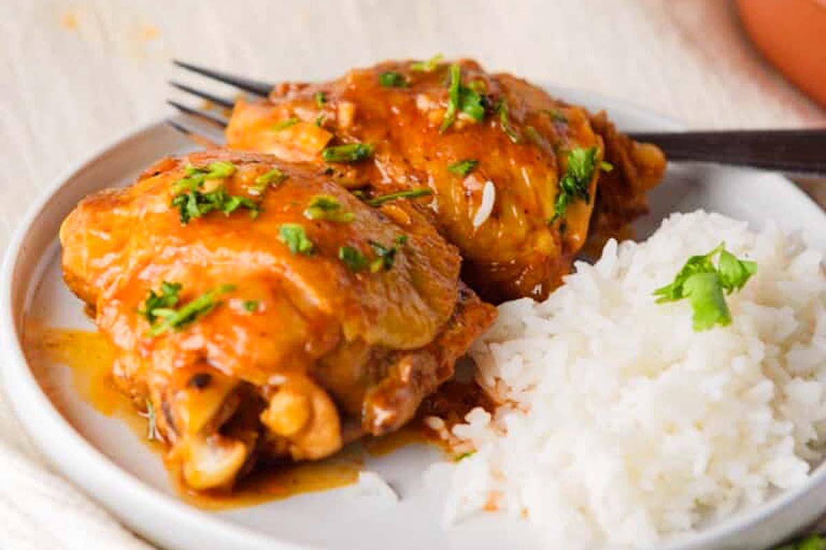 A plate with two pieces of saucy cooked chicken thigh, garnished with chopped herbs, accompanied by a portion of plain white rice. Perfect for those seeking delectable chicken thigh recipes.