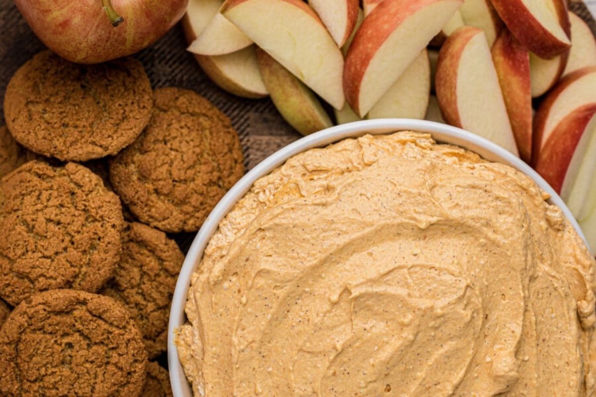 A bowl of creamy dip surrounded by sliced apples and round cookies on a wooden surface.