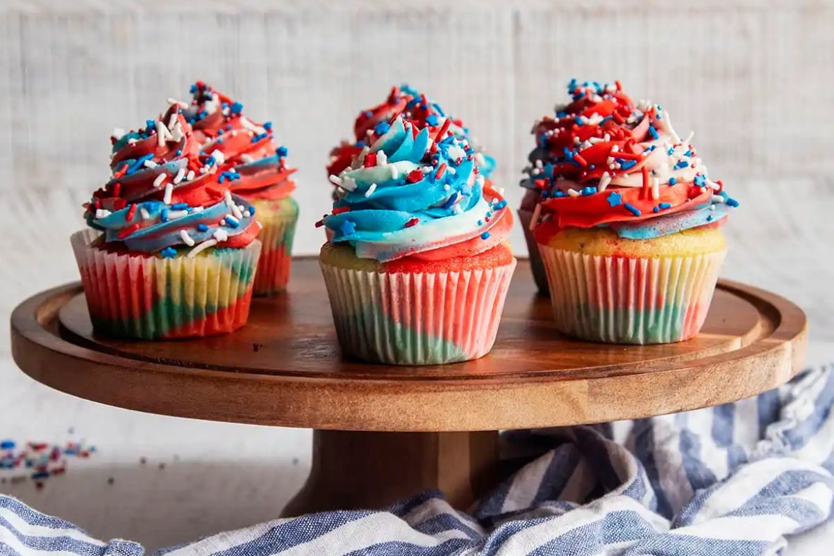 Five colorful cupcakes with multi-colored frosting and sprinkles are displayed on a wooden cake stand. A blue and white cloth, perfect for highlighting these patriotic desserts, is underneath the stand.
