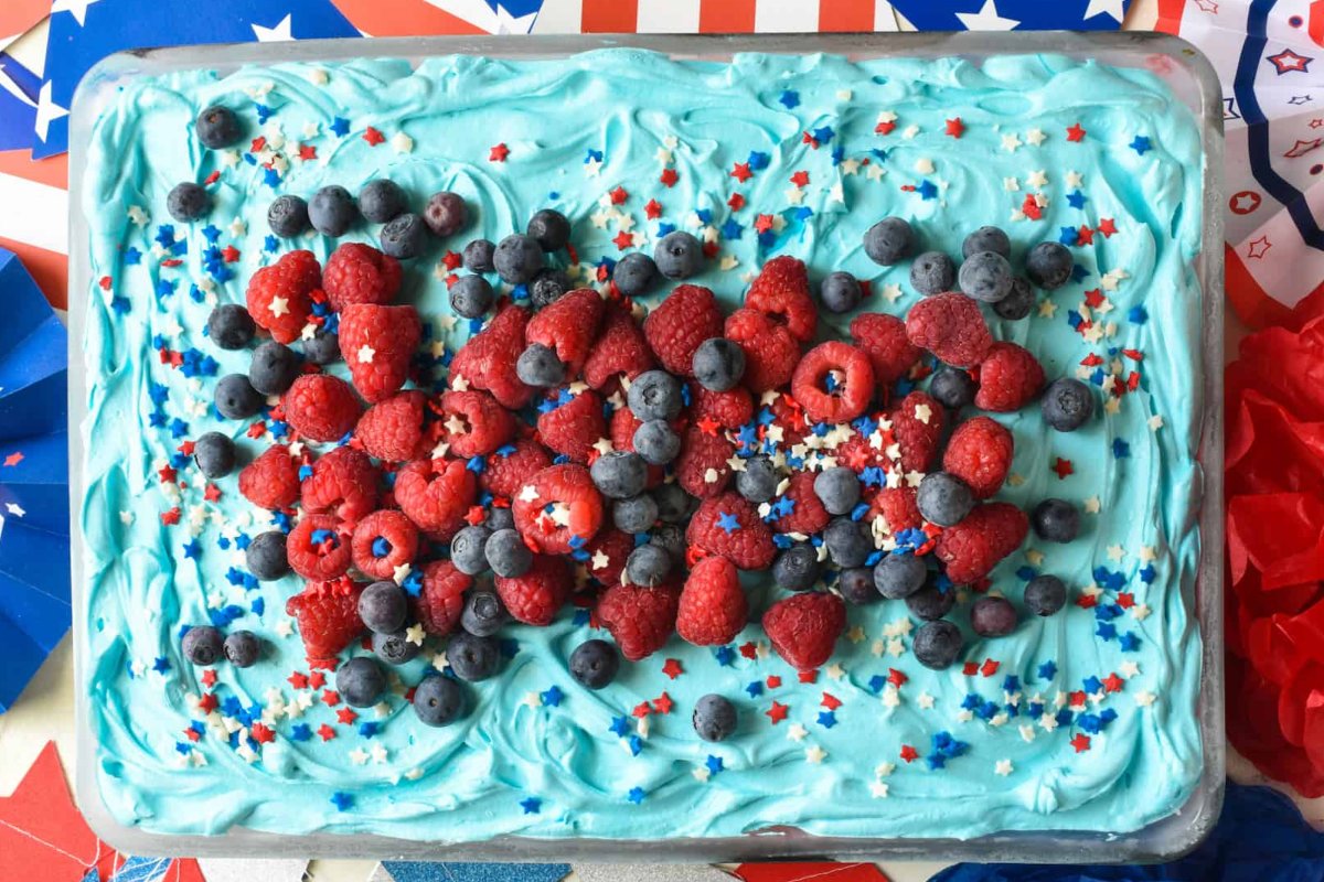 A rectangular cake with light blue frosting, topped with raspberries, blueberries, and star-shaped sprinkles in red, white, and blue colors. The background features red, white, and blue decorations. A perfect choice for those looking for Patriotic Desserts to celebrate in style!