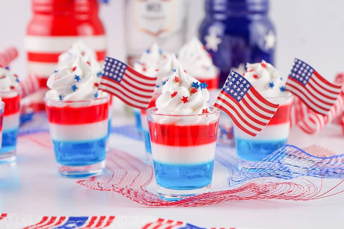 Patriotic desserts featuring small cups with red, white, and blue layers topped with whipped cream, star-shaped sprinkles, and American flags, set on a table adorned with red, white, and blue ribbons.