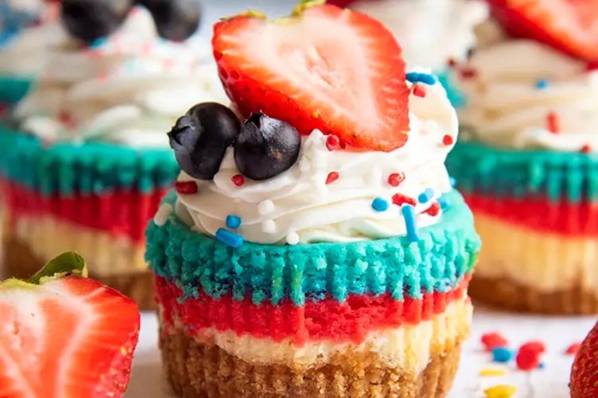 A colorful layered cupcake topped with whipped cream, a slice of strawberry, blueberries, and sprinkles&mdash;perfect for indulging in patriotic desserts.
