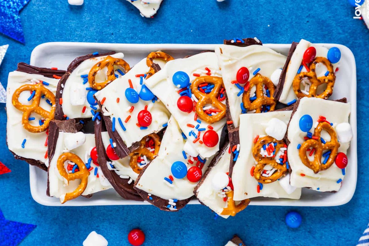 A white tray holds chocolate bark topped with red, white, and blue candy-coated chocolates, pretzels, and sprinkles. This patriotic dessert creation rests on a blue surface scattered with additional candy pieces and sprinkles.