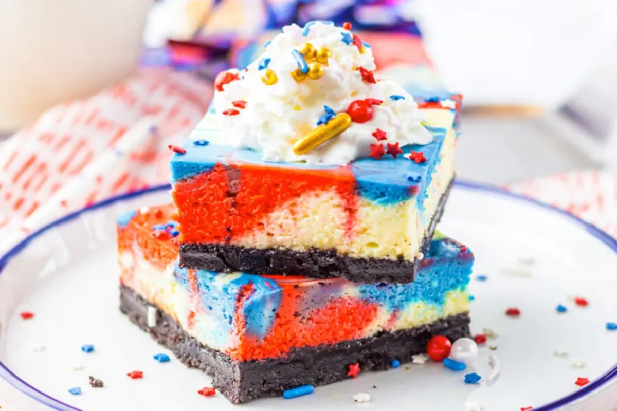 Two colorful cheesecake slices with vibrant red, blue, and yellow swirls, topped with whipped cream and sprinkles, sit on a plate. Perfect as patriotic desserts, they feature a dark chocolate crust for a delightful contrast.