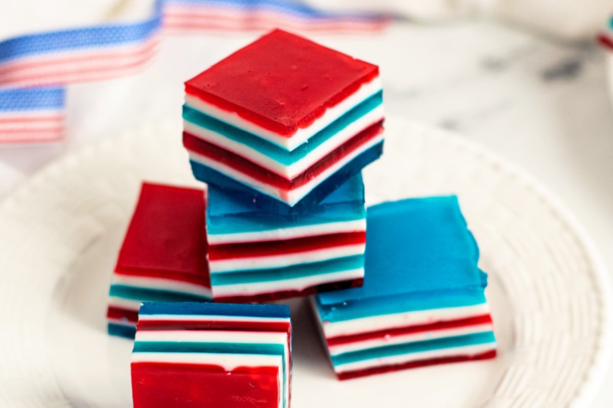 A stack of colorful layered gelatin squares in red, white, and blue sits on a white plate, embodying the essence of patriotic desserts. An American flag-patterned ribbon is blurred in the background.