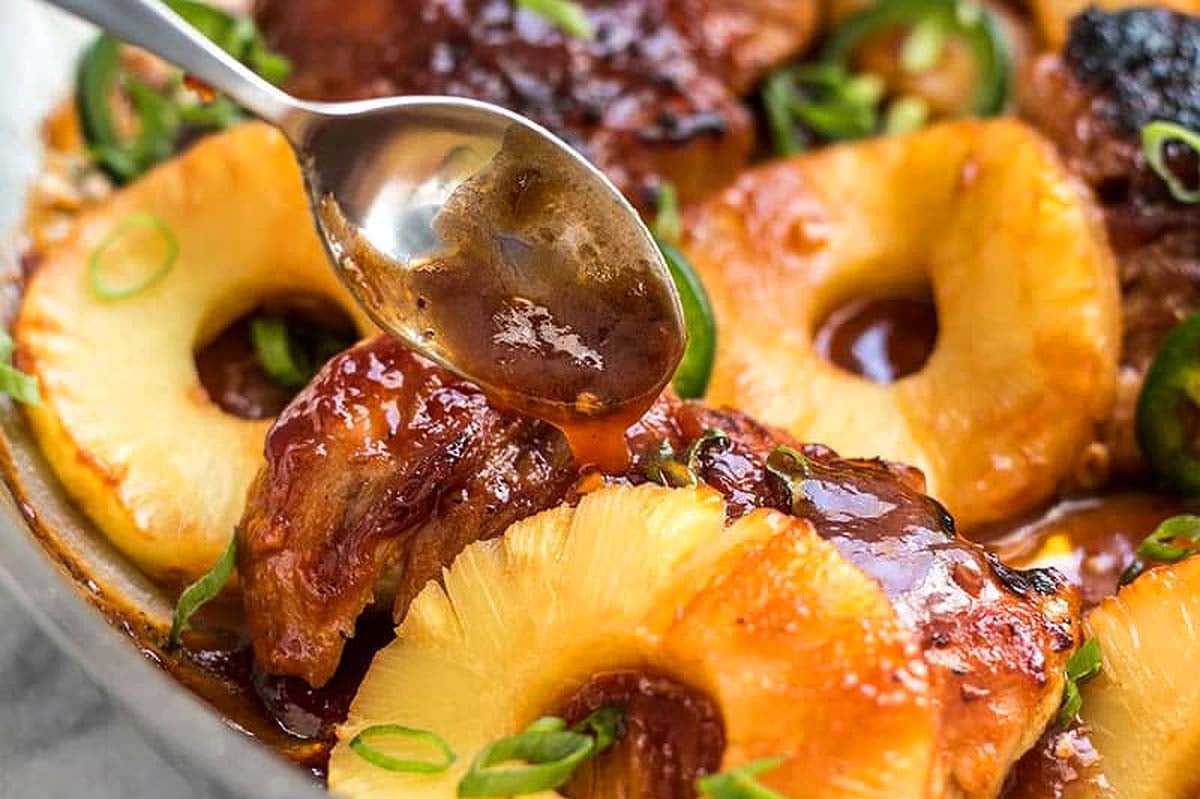 A spoon drizzling glaze over baked chicken thighs topped with pineapple rings and garnished with sliced green onions, making it a delightful addition to your favorite chicken thigh recipes.