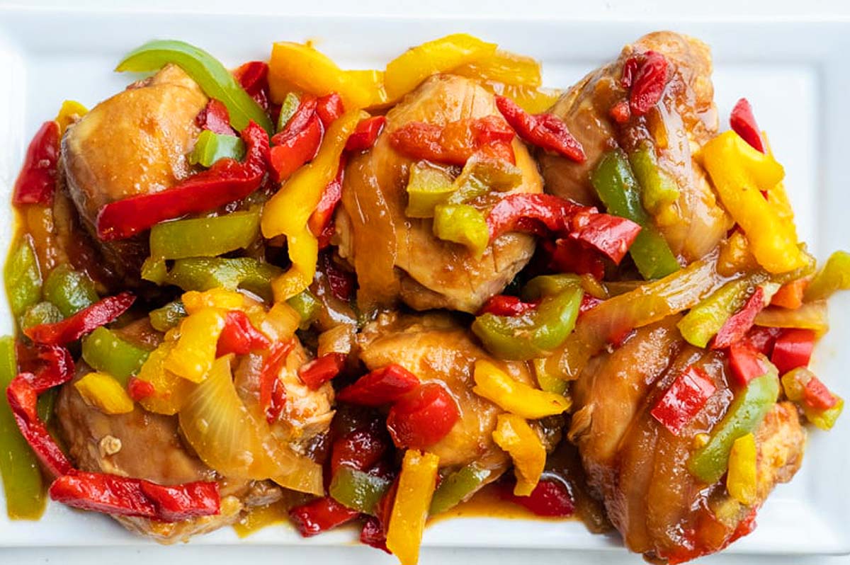 A rectangular plate of cooked chicken thigh pieces topped with mixed sautéed bell peppers and onions in a brown sauce.
