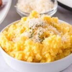 A bowl of slow cooker creamy polenta topped with shredded cheese and herbs, served alongside a plate with a sausage and a small bowl of extra cheese.