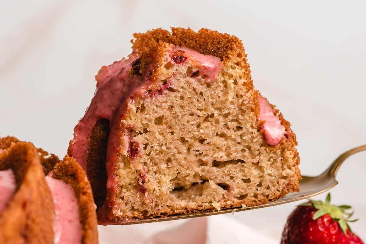 A slice of strawberry Bundt cake with pink strawberry glaze on a serving utensil, next to a fresh strawberry—an ideal treat for those who appreciate shortcut desserts.