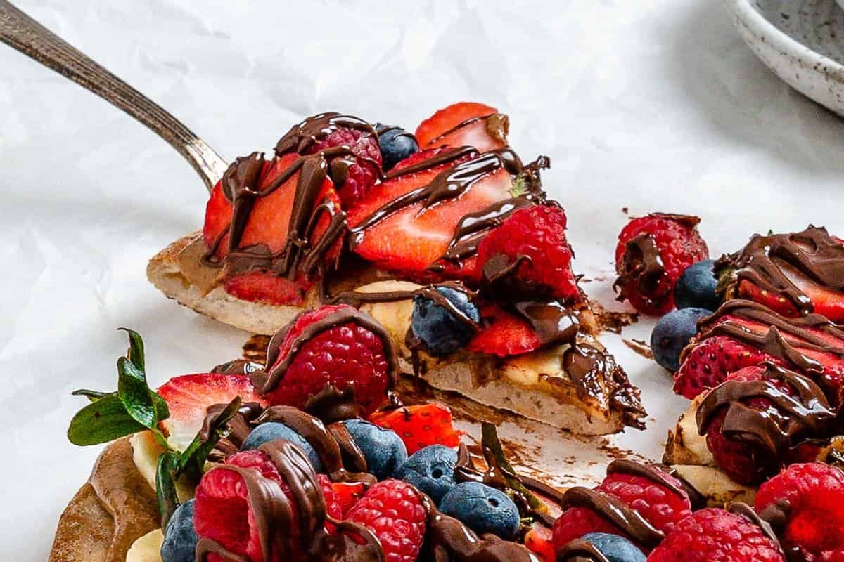 A close-up of an easy fruit dessert pizza topped with strawberries, blueberries, raspberries, banana slices, and drizzled with chocolate. A spatula is lifting one slice.