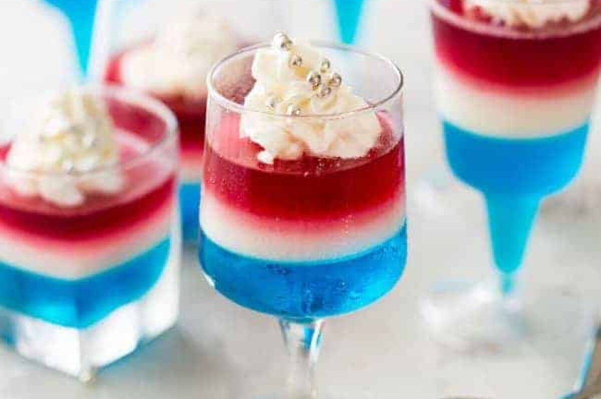 Patriotic desserts featuring layered red, white, and blue in glass cups, topped with whipped cream and small silver balls.