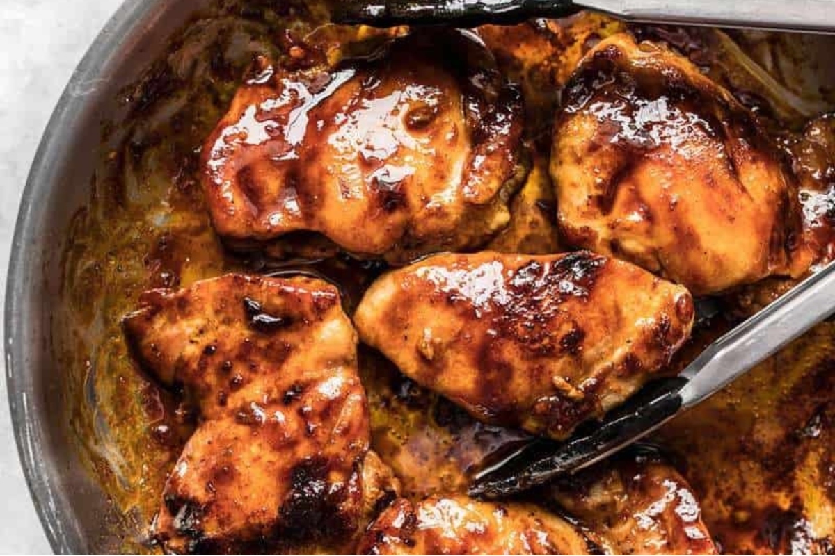 A close-up of glazed and cooked chicken thigh recipes in a pan, with metal tongs resting on top of the chicken.
