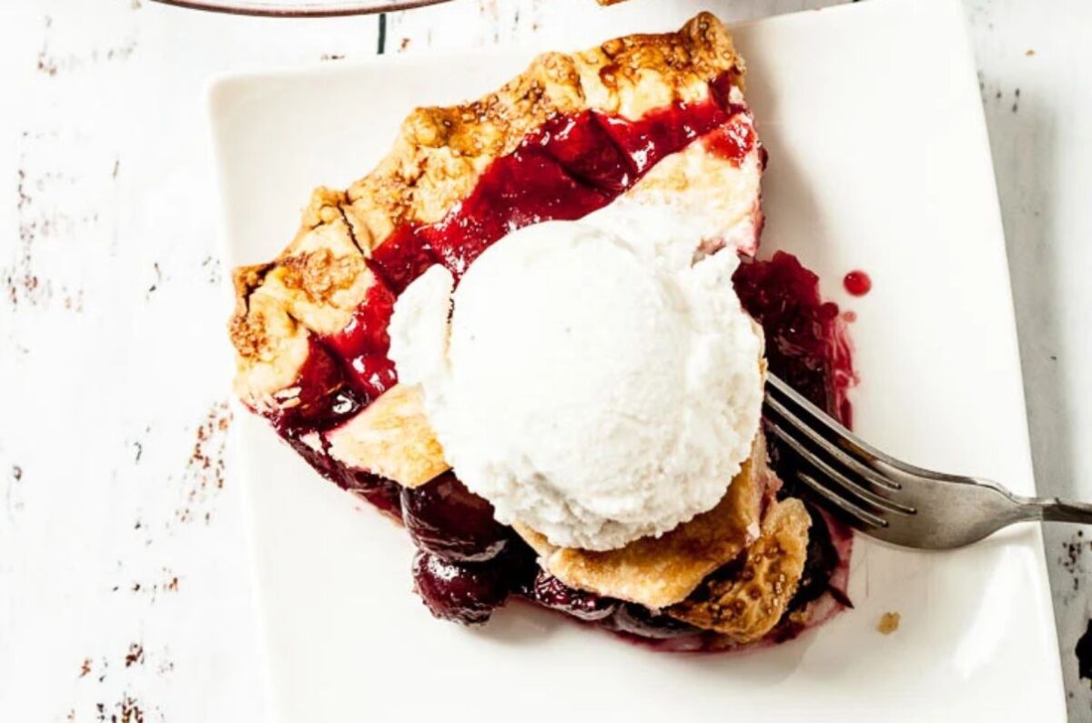 A slice of cherry pie with a lattice crust topped with a scoop of vanilla ice cream on a white square plate, showcasing one of the finest cherry recipes. A fork rests beside the slice.