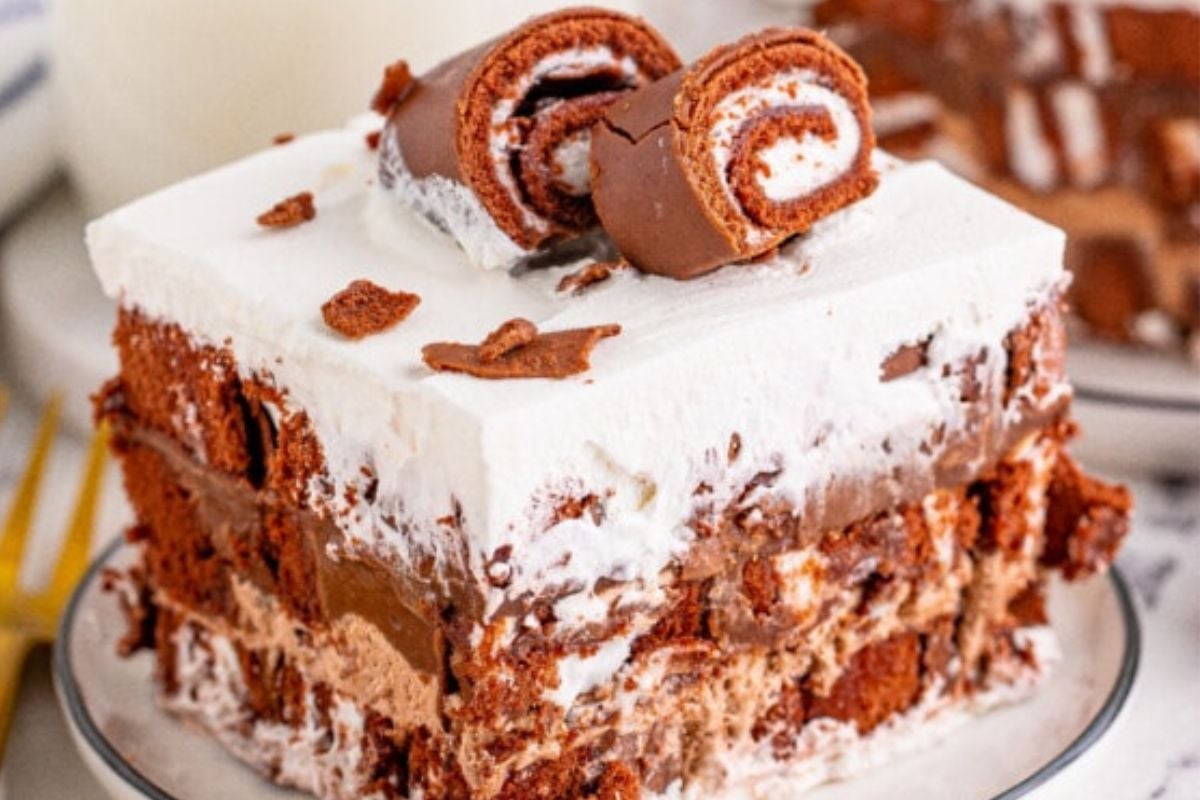 A slice of layered chocolate and cream dessert topped with two small rolled chocolate cakes on a white plate with a fork nearby.