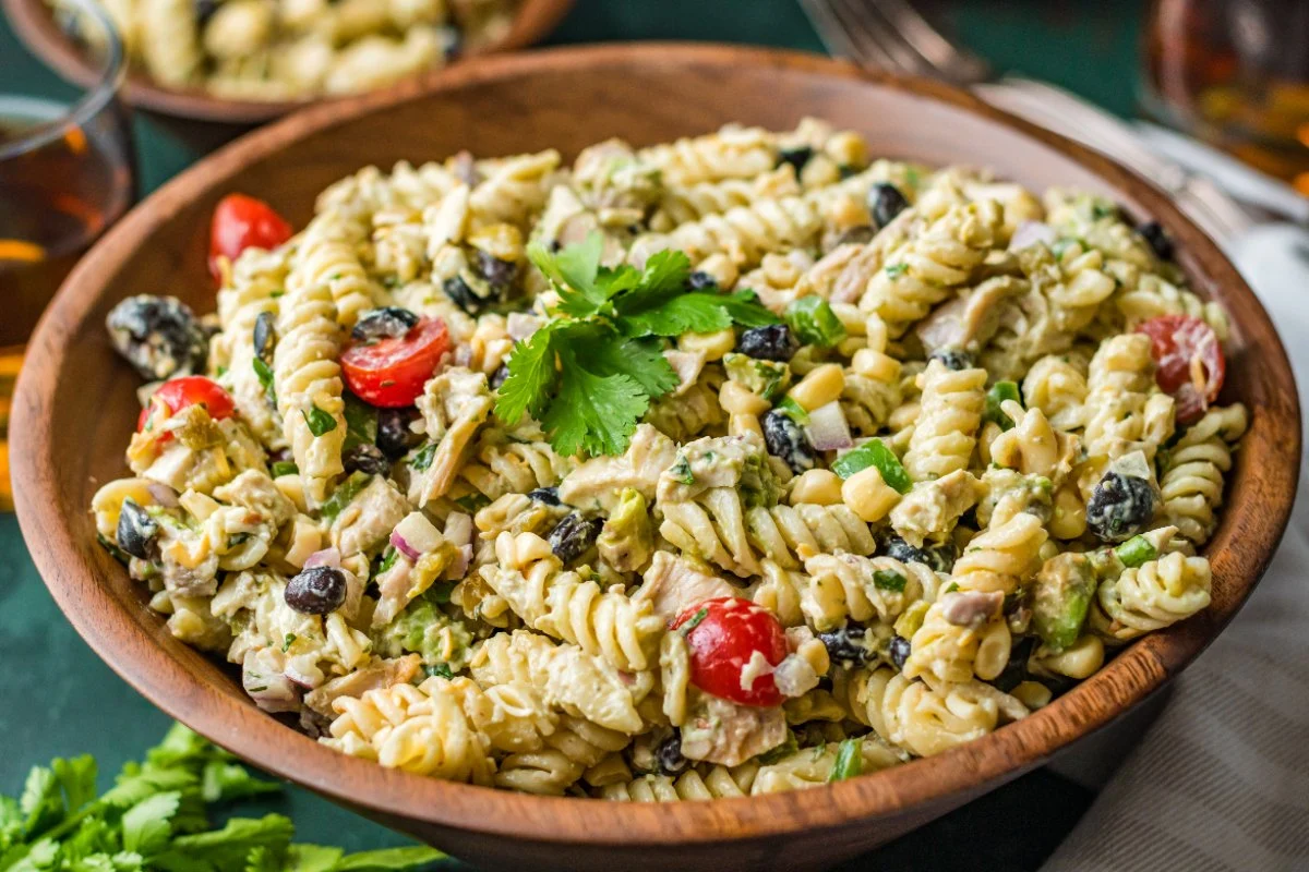 A wooden bowl filled with creamy pasta salad, cherry tomatoes, olives, and fresh parsley garnish.