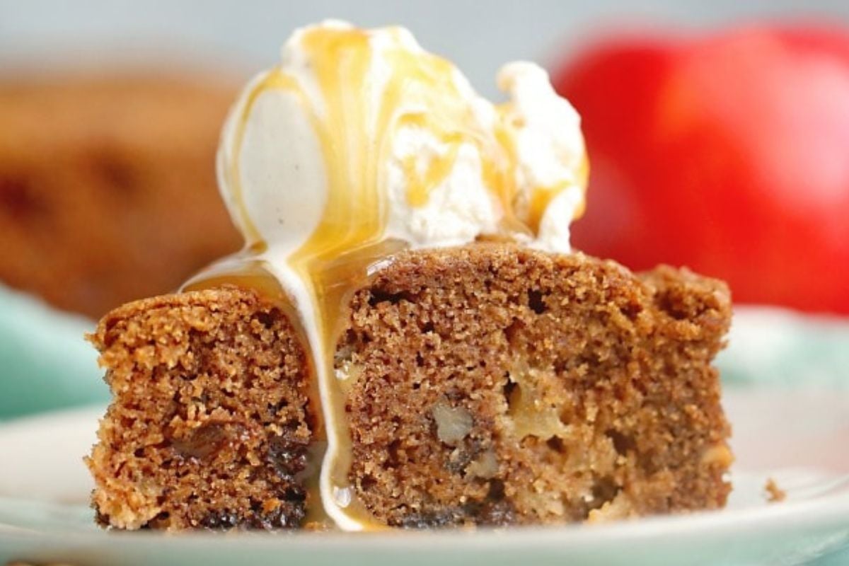 A slice of brown cake topped with a scoop of vanilla ice cream and drizzled with caramel sauce, with an apple in the background.