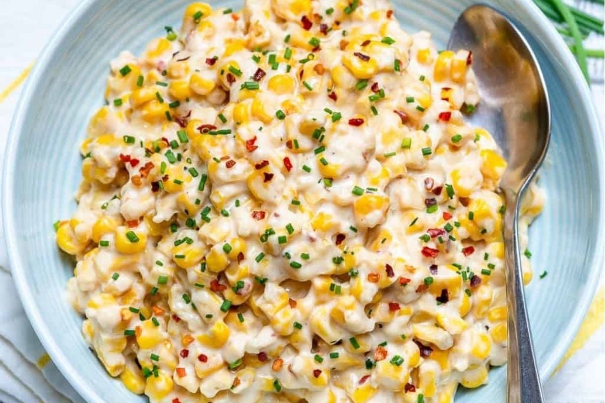 A bowl of creamy corn salad garnished with red pepper flakes and chives, featured in a church cookbook, served on a blue plate with a spoon on the side.