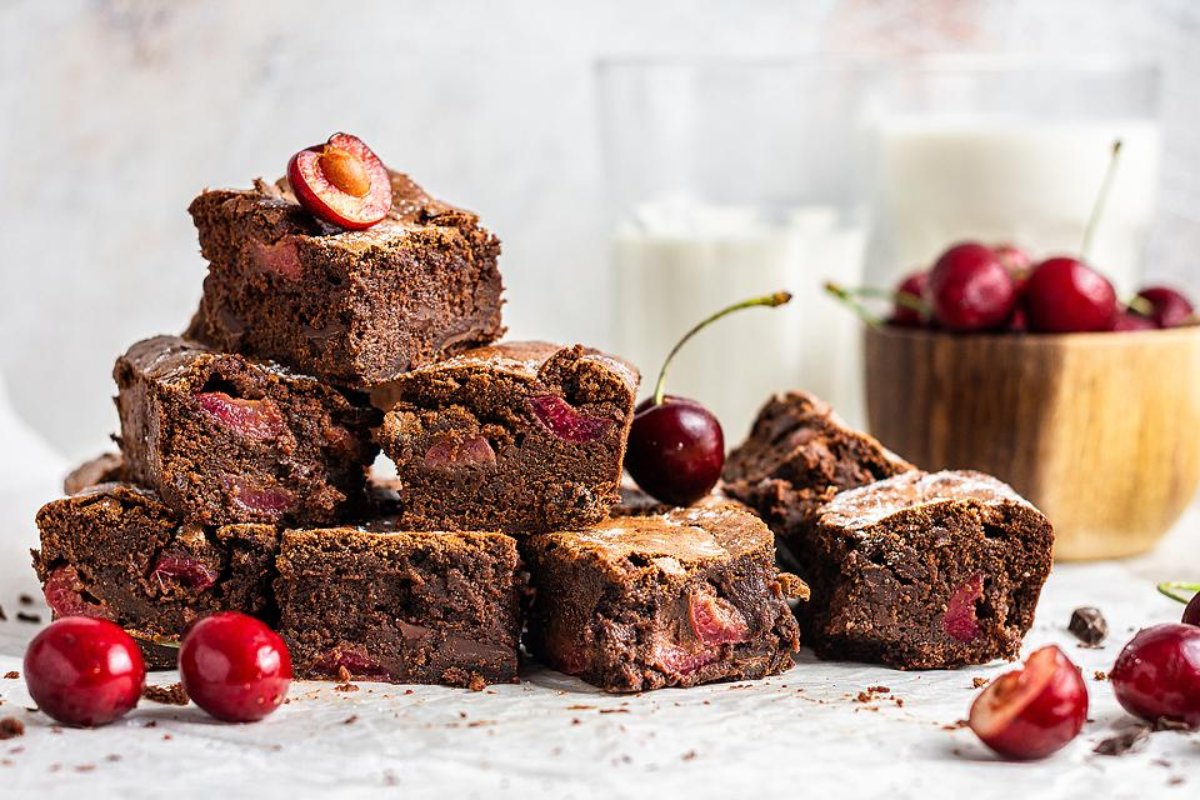 A stack of chocolate brownies with cherries, some whole cherries placed on top and around, a wooden bowl of cherries, and two glasses of milk in the background—a delightful display perfect for cherry recipes.